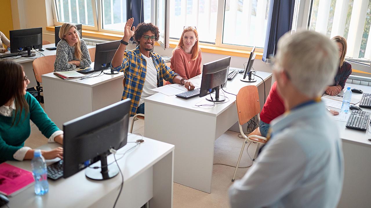 Student at a computer in a classroom raises hand to ask a question to the instructor